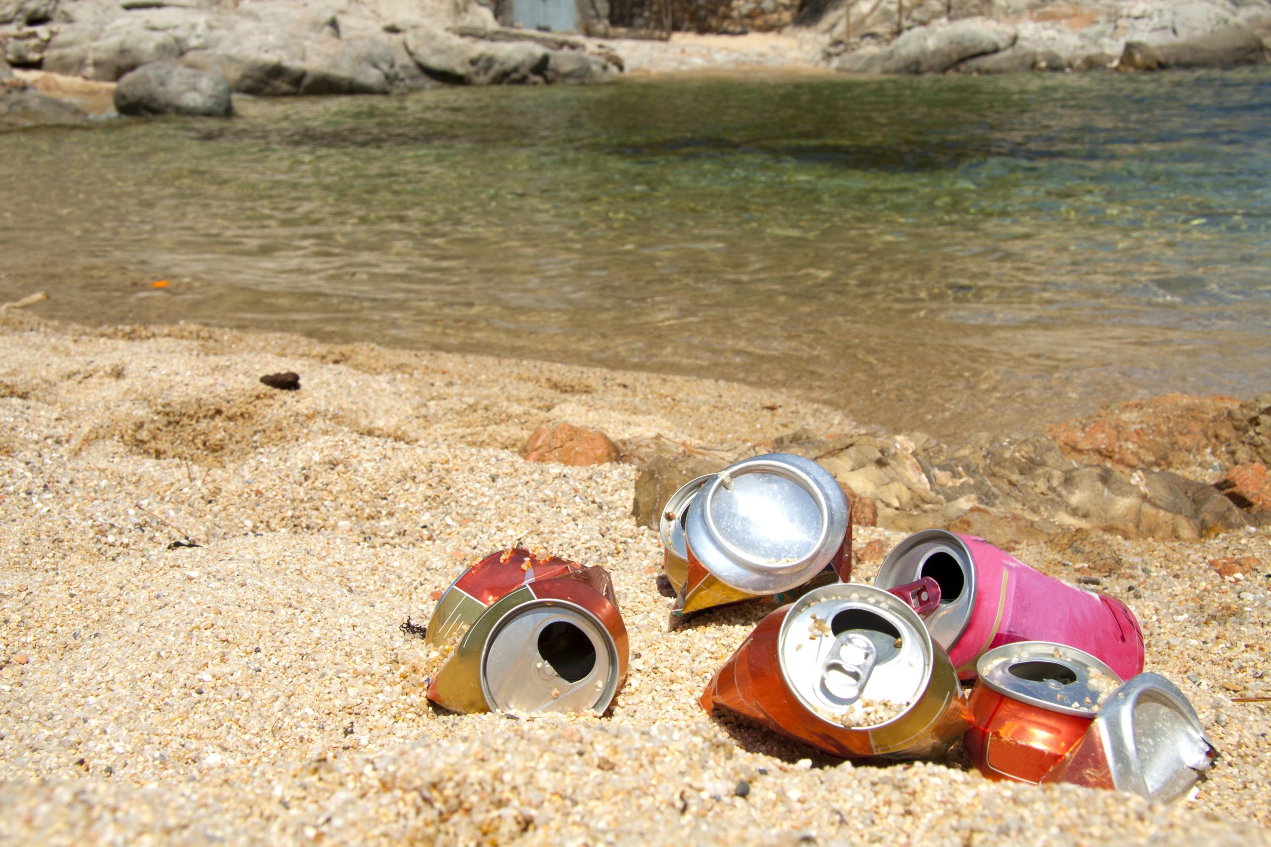 Crushed soda cans litter a sandy beach near clear, calm water. Rocks line the shoreline, highlighting environmental pollution concerns.